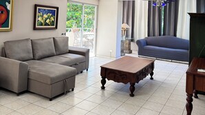 Un salon avec un canapé gris, une table basse en bois et un canapé bleu. Deux œuvres d’art encadrées ornent le mur, et une fenêtre est agrémentée de rideaux.