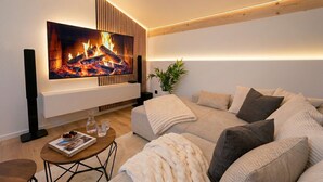 Een moderne woonkamer met een grote flatscreen televisie aan de muur, een beige bank met meerdere kussens, een klein bijzettafeltje met een tijdschrift en glazen, en een potplant.