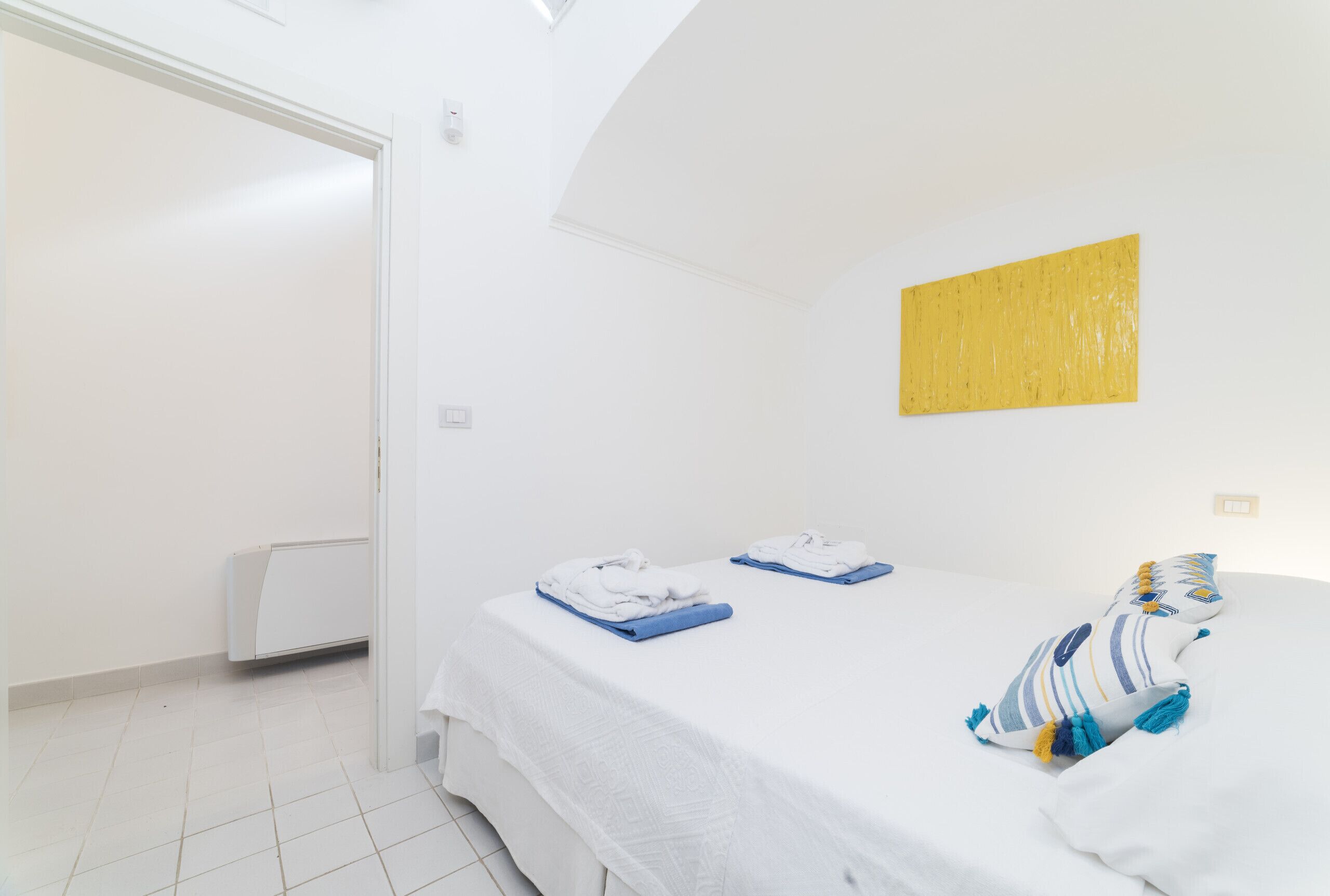 Ein sauberes, weißes Zimmer mit einem Bett, einem Heizkörper und einem gelben Kunstwerk an der Wand.