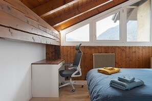 Un dormitorio con techo de madera, un escritorio, una silla, una cama con ropa de cama azul y vistas a las montañas a través de una ventana.
