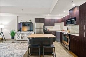Eine moderne Küche mit dunklen Holzschränken, Edelstahlgeräten, einem Holztisch mit zwei Stühlen und einem kleinen Fernseher auf einem weißen Schrank.