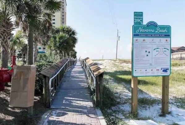 A pathway leading to a beach area with a sign indicating 'Navarre Beach' and a temperature of 39°C. - Sand Dollar Quarters - 3BR, Gulf Access, Sound Dock, Hot Tub, Community Pool (Navarre)