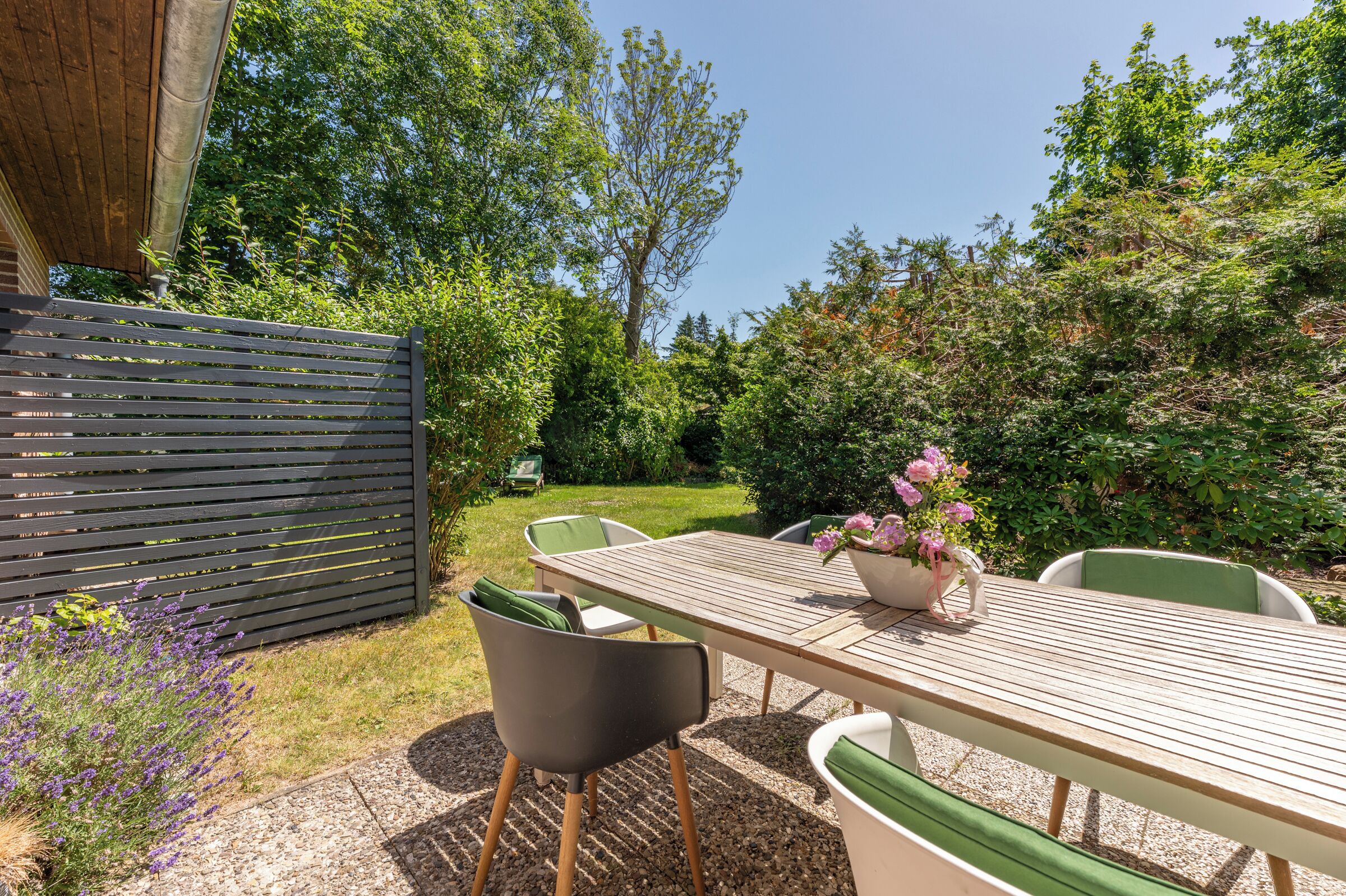 Eine Terrasse im Freien mit Tisch und Stühlen, umgeben von viel Grün und einem Holzzaun.