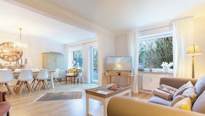 Ein geräumiges Wohnzimmer mit einer Couch, einem Couchtisch und einem Fernseher. Es gibt einen Essbereich mit Stühlen und einem Tisch, sowie ein Fenster mit Vorhängen.