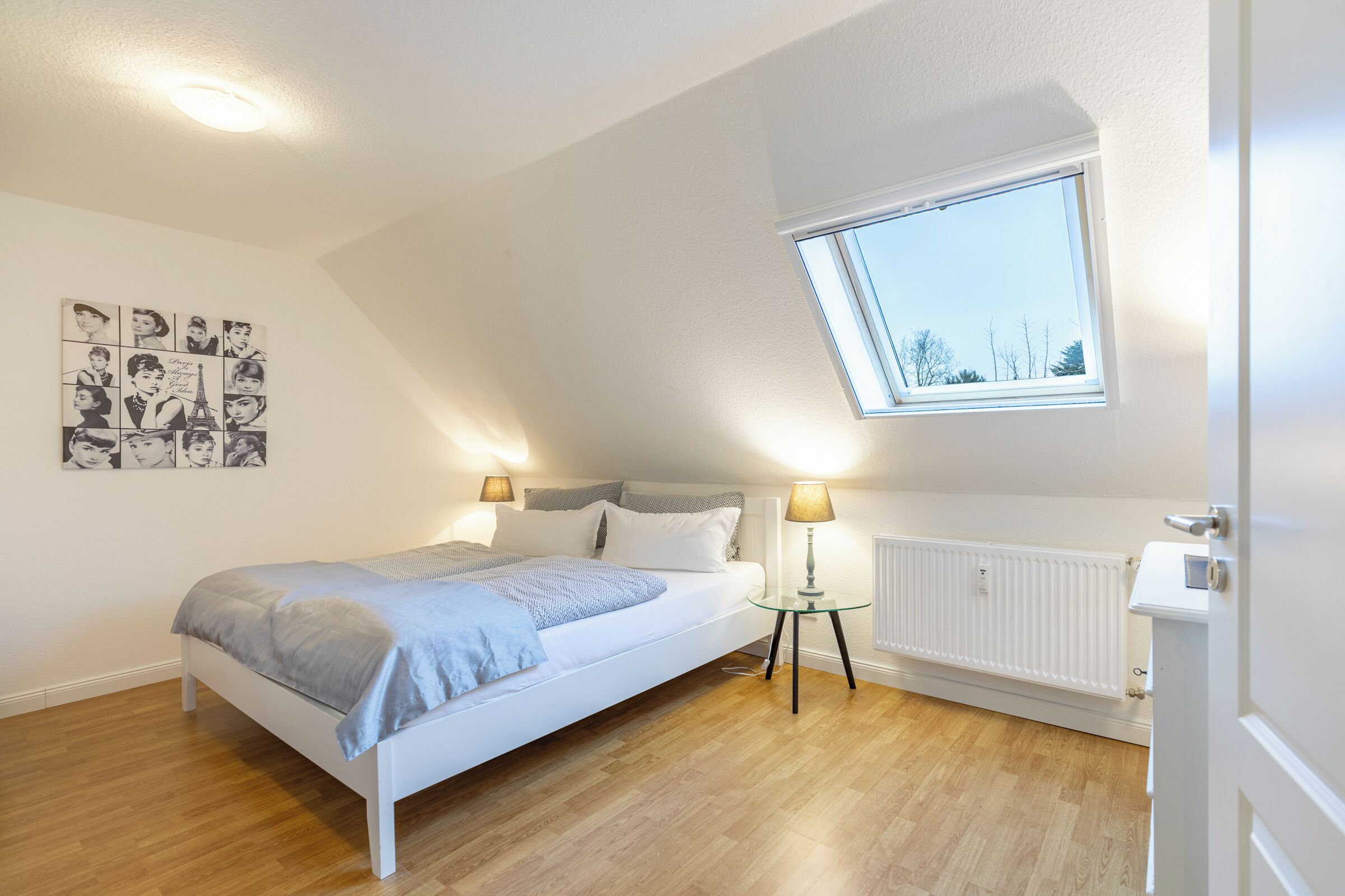 Ein Schlafzimmer mit schrägem Dach, einem Oberlicht, einem Bett mit grauer Decke, zwei Nachttischlampen, einem Heizkörper und einer Wand mit einer schwarz-weißen Fotocollage.