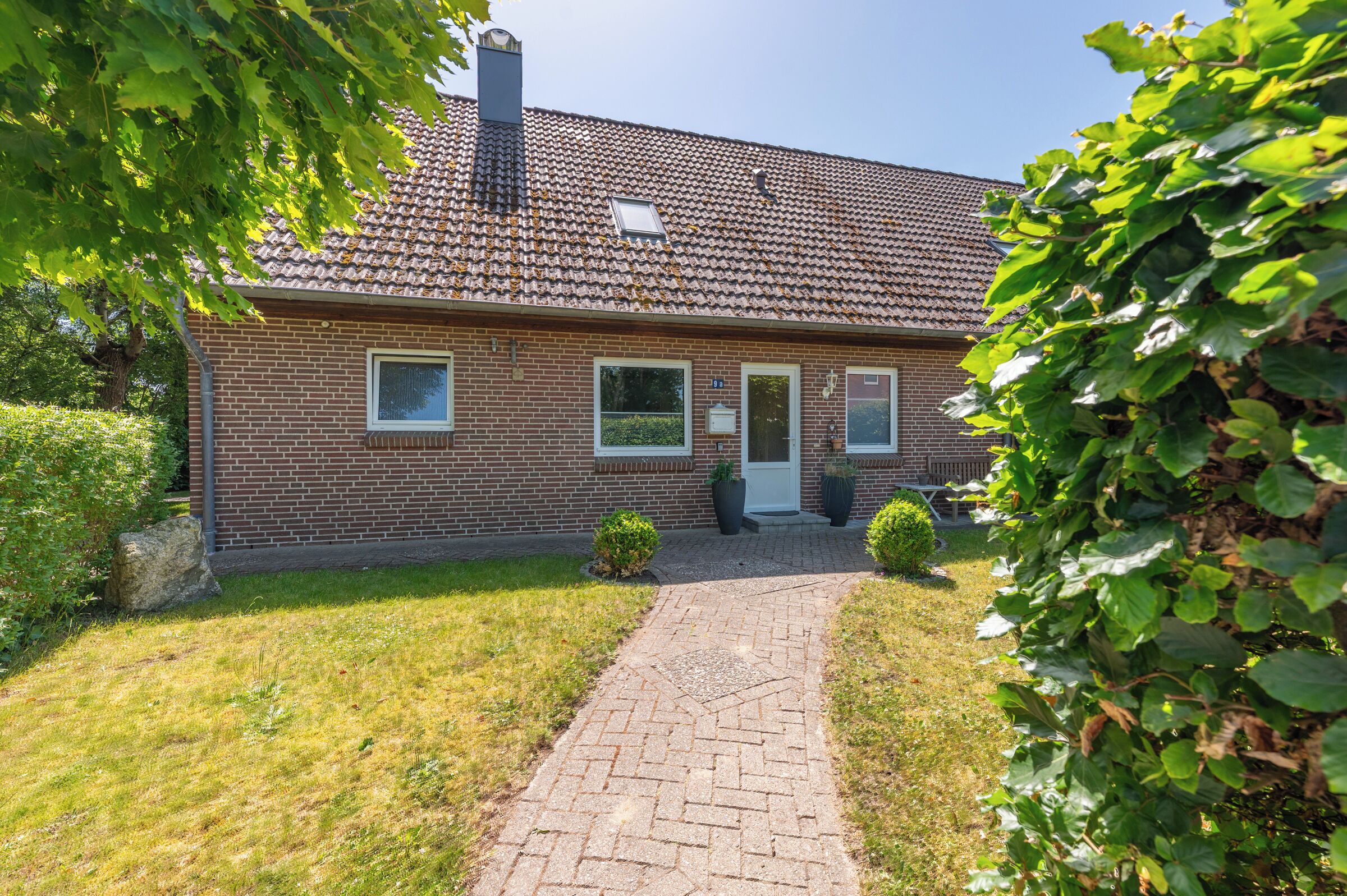 A brick house with a tiled roof, a paved driveway, and a small front yard with a bush and a potted plant.