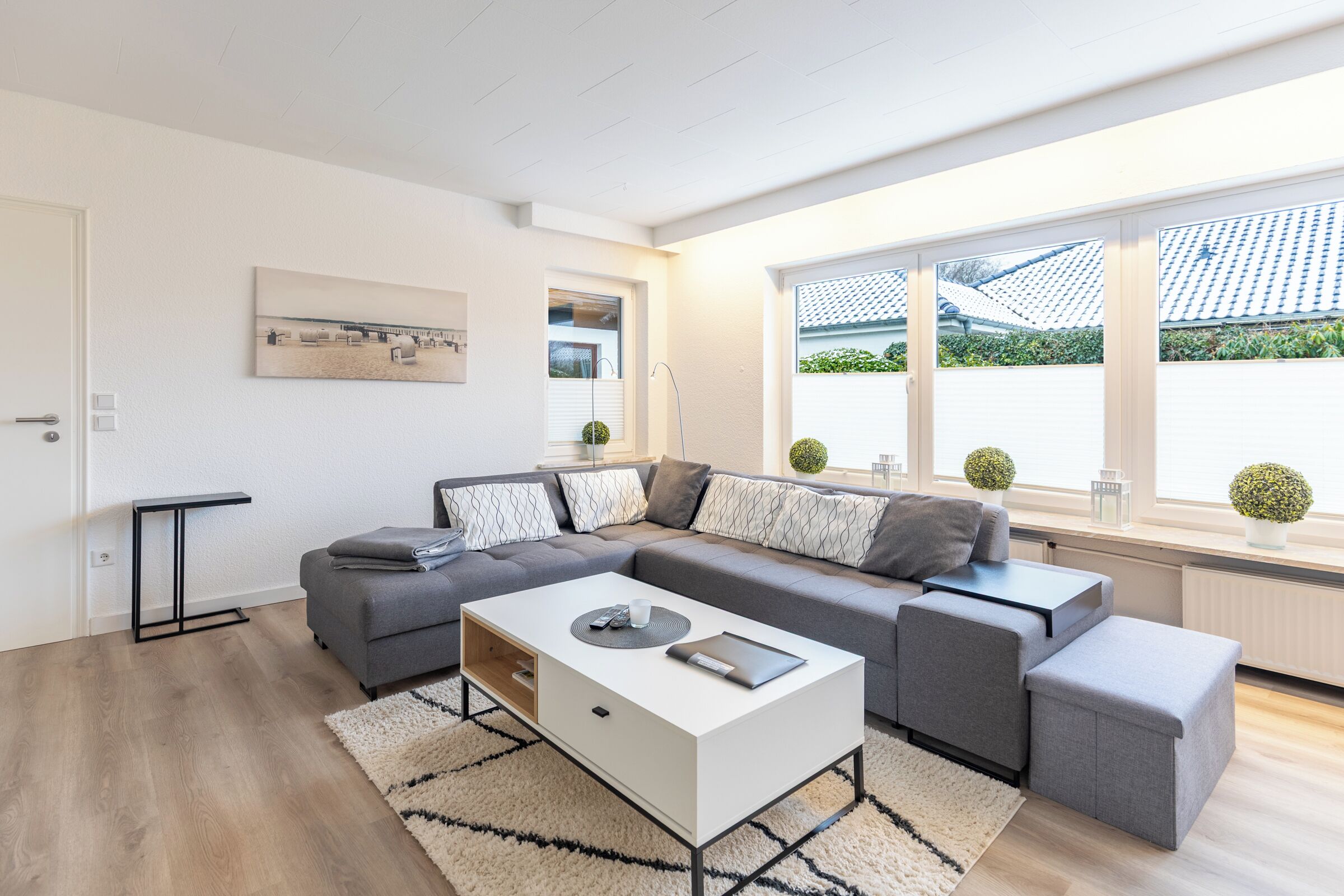 A modern living room with a grey sectional sofa, a white coffee table, and a black side table. There is a large window with a view of a tiled roof and greenery.