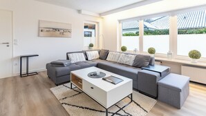 Una sala de estar moderna con un sofá seccional gris, una mesa de centro blanca y una mesita de noche negra. Hay un ventanal con vista a un tejado de baldosas y follaje.