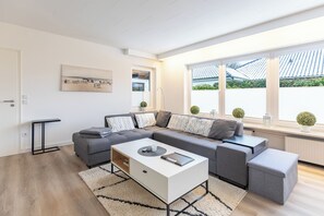 Ein modernes Wohnzimmer mit einem grauen Ecksofa, einem weißen Couchtisch und einem schwarzen Beistelltisch. Ein großes Fenster bietet Blick auf ein Ziegeldach und Grünflächen.