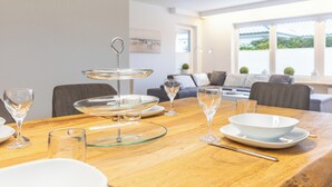 Una mesa de comedor decorada con cristalería transparente y platos, un centro de mesa de vidrio escalonado, y una vista de una sala de estar moderna con un sofá y ventanas.