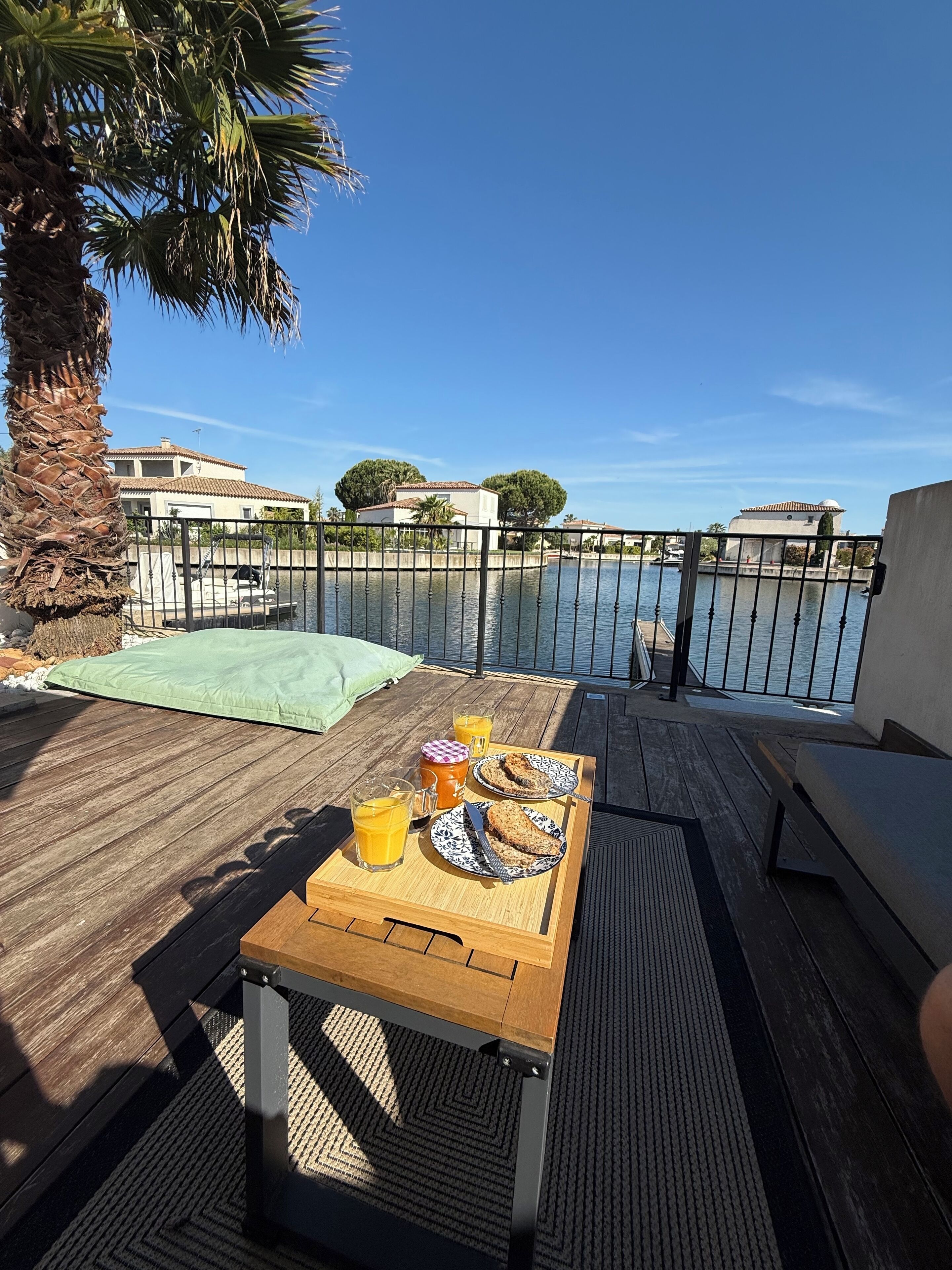 A wooden deck with a table set for breakfast, overlooking a body of water and a residential area.