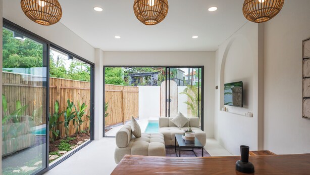 A modern living room with a wooden dining table, a sofa set, and a large window overlooking a garden. - Bohemia Villa by Betterplace (Canggu)