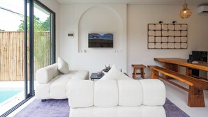 A modern living room with a large sofa, a wooden bench, a TV mounted on the wall, and a view of the outdoors. - Bohemia Villa by Betterplace (Canggu)