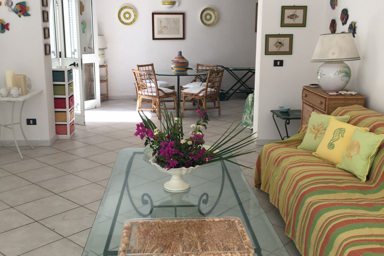 Un soggiorno con un tavolino in vetro, un divano con cuscini a righe, un'area pranzo con tavolo e sedie, e una parete decorata con quadri e piatti decorativi.