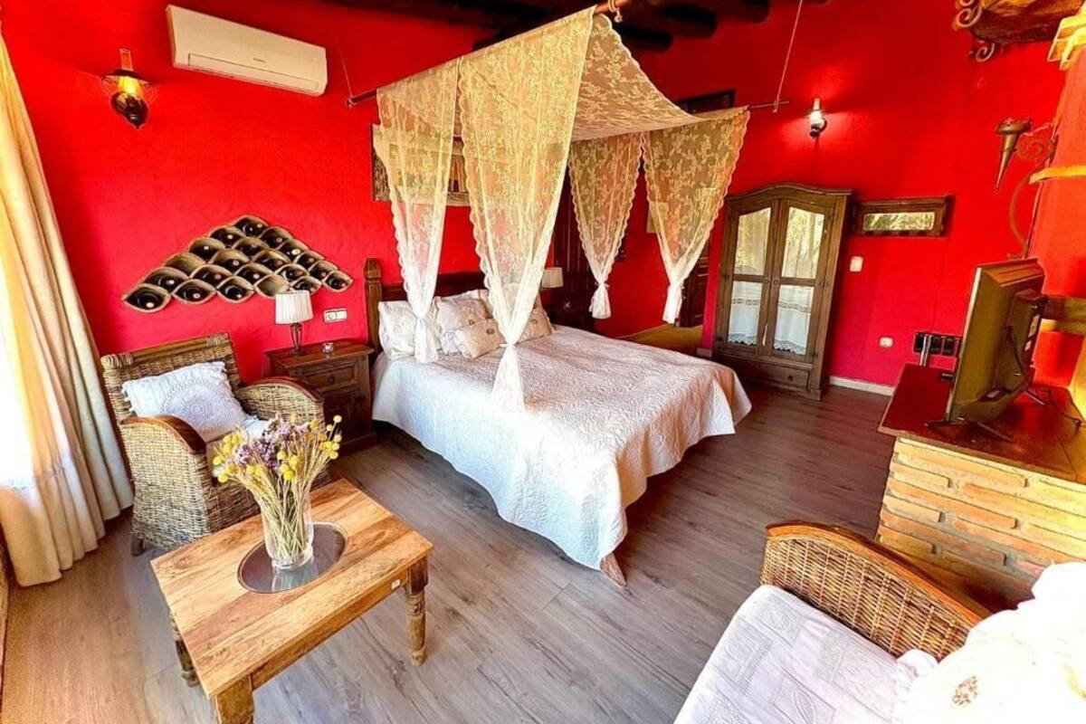 Une pièce avec un mur rouge, un lit à baldaquin, des meubles en osier et une table en bois ornée d’un vase de fleurs.