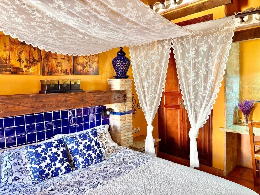 Een slaapkamer met een bed, een houten hoofdbord, een deken met blauw-wit patroon, een wit baldakijn, een blauwe vaas en een muur met een mozaïek ontwerp.