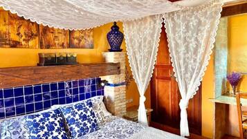 Een slaapkamer met een bed, een houten hoofdbord, een deken met blauw-wit patroon, een wit baldakijn, een blauwe vaas en een muur met een mozaïek ontwerp.