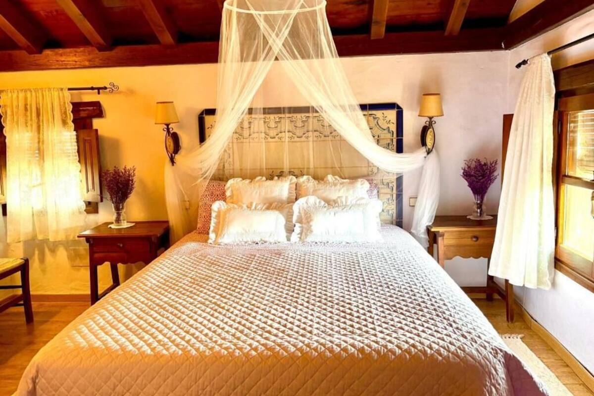 Une chambre à coucher avec un grand lit, un baldaquin, deux tables de chevet avec des lampes, une petite table et une fenêtre avec des rideaux.
