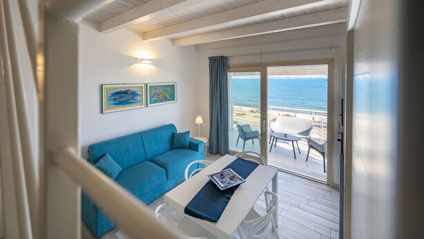 A room with a blue sofa, a dining table with a blue table runner, and a view of the beach through a glass door. - Apartment Lights (Avola)