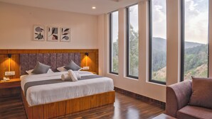 A modern bedroom with a large bed, wooden headboard, bedside lamps, and a view of the outdoors through large windows. - Hardys Retreat (Shimla)