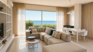 A modern living room with a large L-shaped sofa, a round coffee table, a dining area with a white table and chairs, and a view of the ocean.