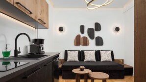 A modern kitchen with a black countertop, a coffee machine, and a green glass container. There is a black sofa with patterned pillows and a small wooden table with a bowl. - Voukourestiou Signature Suite (Athens)