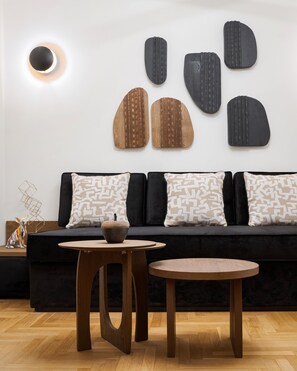Un salon moderne avec un canapé noir, des coussins à motifs, une table ronde en bois sur laquelle est posé un bol, et des œuvres d'art abstraites accrochées au mur.