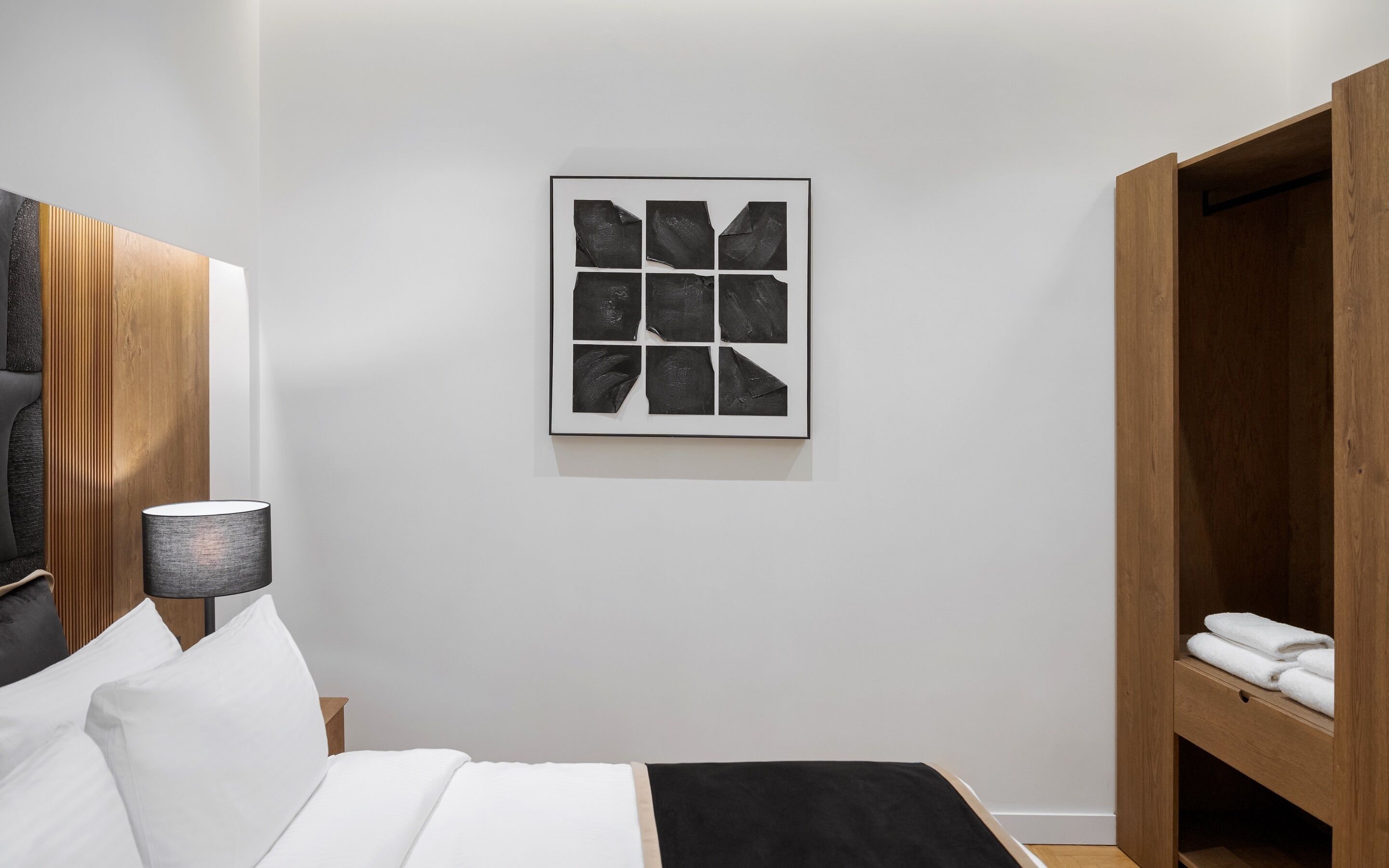 Un dormitorio moderno con una cama, una mesita de noche, una lámpara, un armario de madera y una obra de arte abstracta enmarcada en la pared.