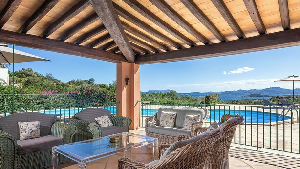 A covered outdoor seating area with wicker furniture, a glass table, and a view of a swimming pool and mountains. - Classic villa, Cala di Volpe, minutes away from Sardinia's most iconic beaches (Sardegna)