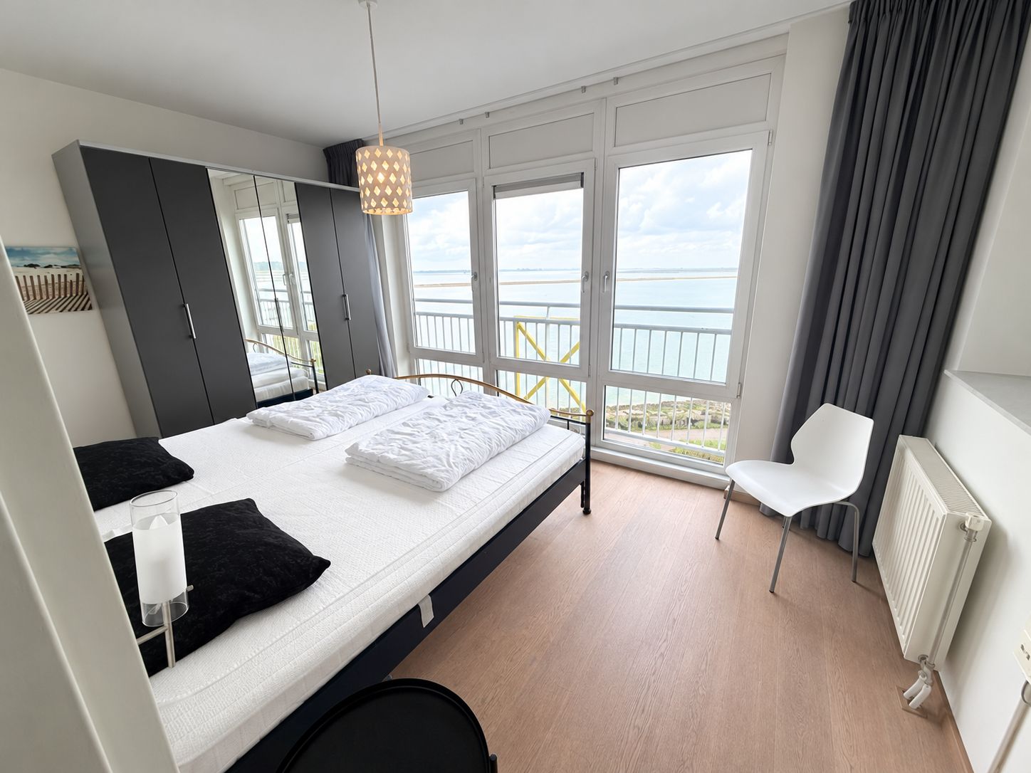 A modern bedroom with a large bed, a chair, a radiator, and a view of the sea through large windows.