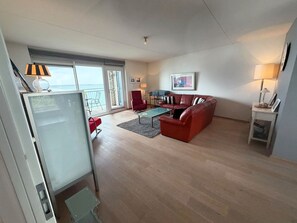 Ein Wohnzimmer mit einem roten Sofa, einem Glastisch und einem roten Sessel. Ein Balkon bietet Blick auf das Meer.