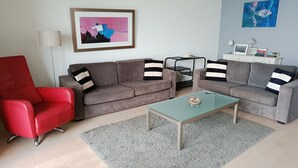 Ett vardagsrum med en röd fåtölj, två grå soffor med randiga kuddar, ett glasbord, en grå matta, en inramad tavla och ett litet vitt bord med böcker och en lampa.