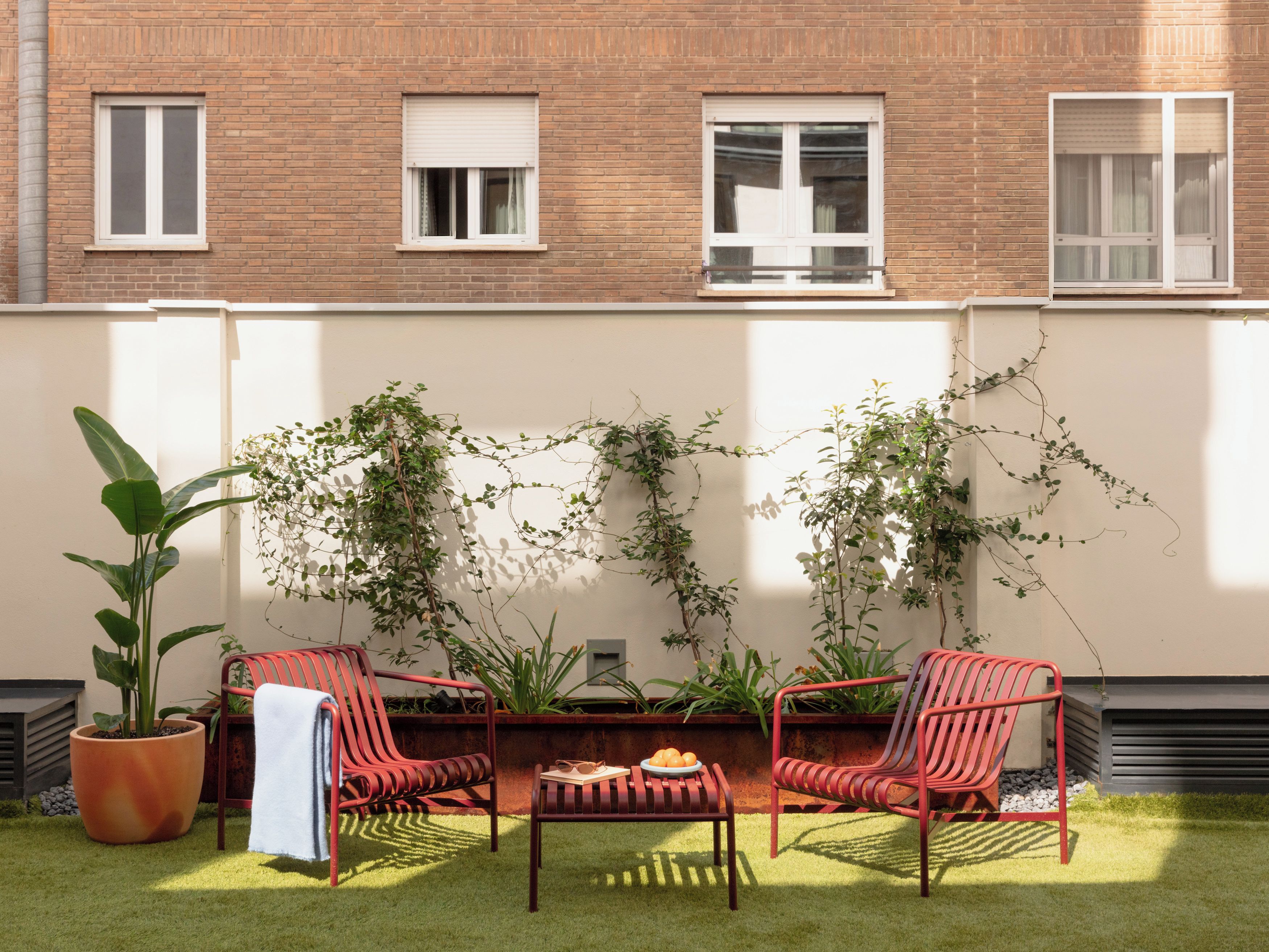 Un patio con sillas rojas, una mesa y plantas en macetas frente a una pared de ladrillo con ventanas.