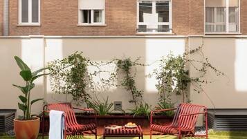 Un patio con sillas rojas, una mesa y plantas en macetas frente a una pared de ladrillo con ventanas.