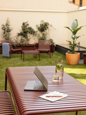 Une terrasse avec une table, un ordinateur portable, un verre d’eau et une plante en pot.
