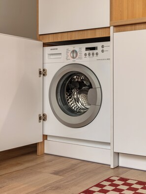 A front-loading washing machine in a kitchen with wooden cabinets and a checkered rug. - Numa Madrid Atocha (Madrid)