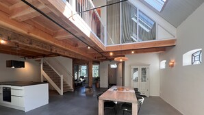 Ein großzügiges Loft mit hohen Decken, freiliegenden Holzbalken, einem Essbereich mit langem Tisch und Stühlen, einer Küche mit Einbauofen und einer Treppe, die zu einer oberen Ebene führt.