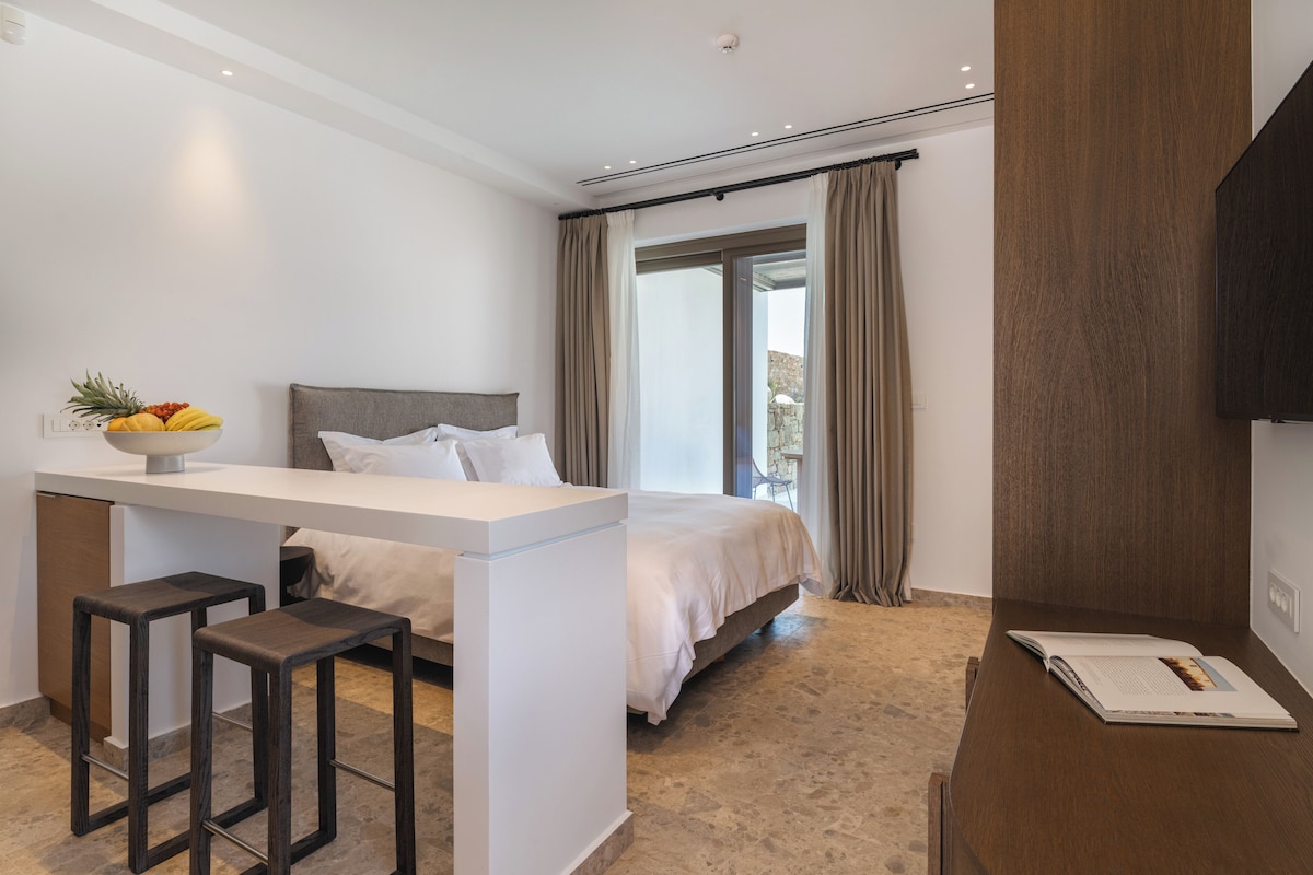 Quarto de hotel moderno com uma cama, uma pequena mesa com banquetas, vista para o exterior e uma grande janela com cortinas.