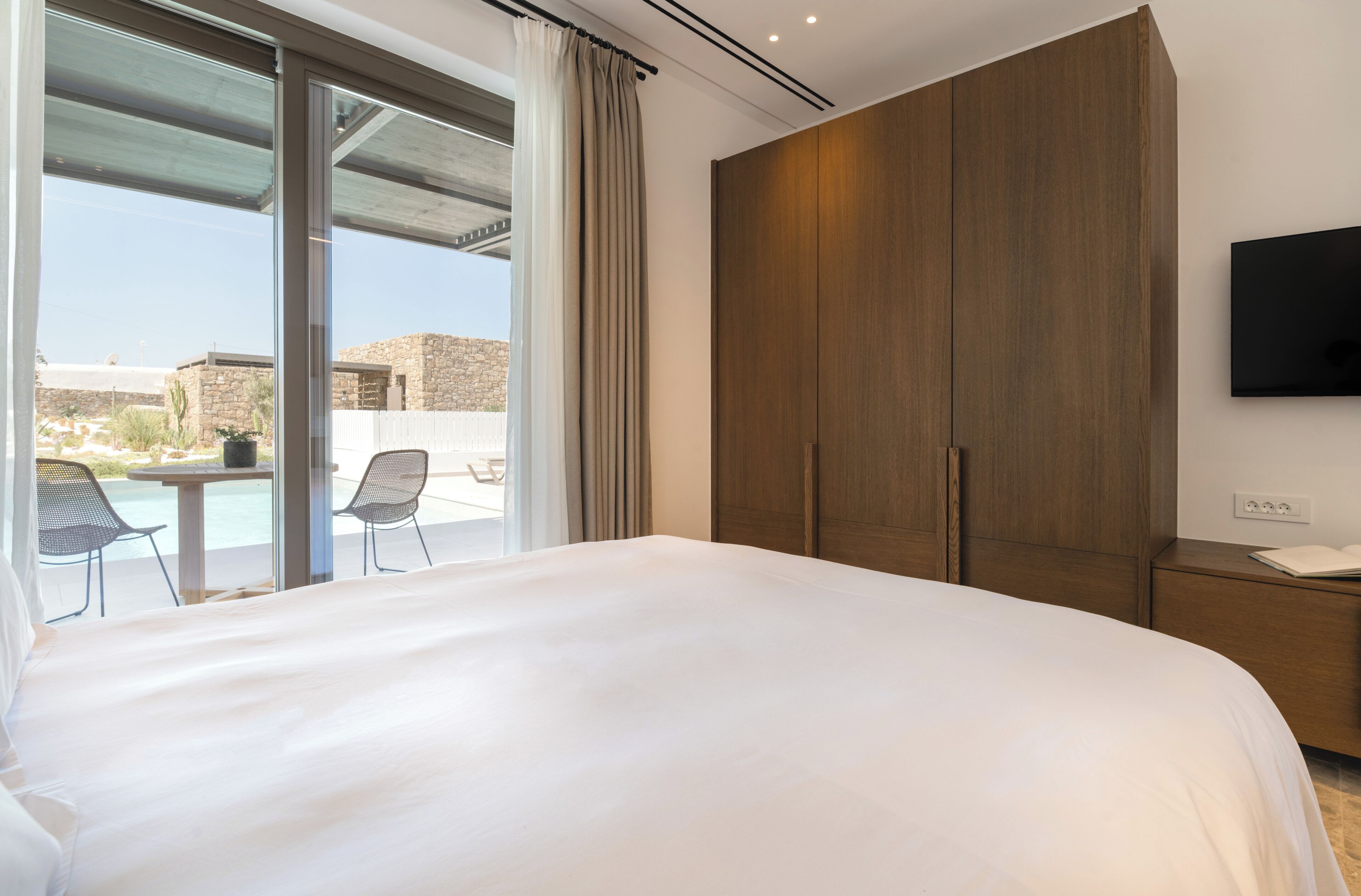 A hotel room with a large bed, a TV mounted on the wall, and a sliding glass door leading to an outdoor area with a table and chairs.