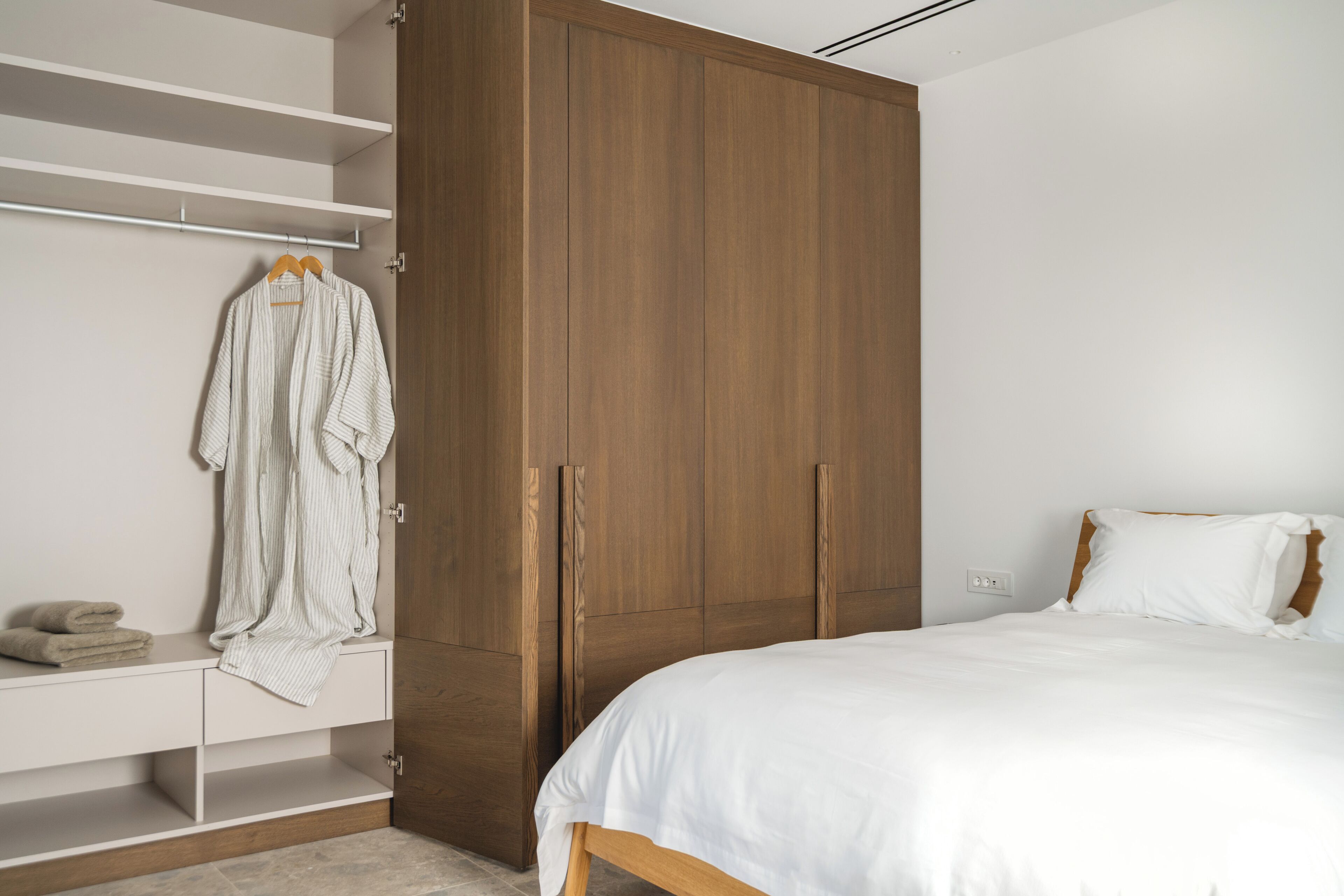 Een slaapkamer met een bed, een houten garderobe en een witte nachtkastje met handdoek en badjas.