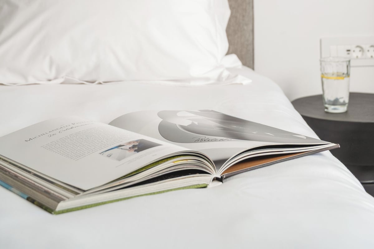Cama bem arrumada com revista, um copo de água e mesa de cabeceira com abajur.