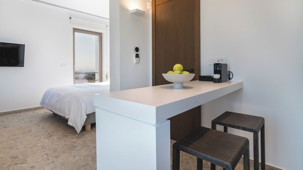 A modern hotel room with a bed, a small kitchenette, a bowl of fruit on the counter, and a TV mounted on the wall. - Omvrodektis Village (Mykonos)