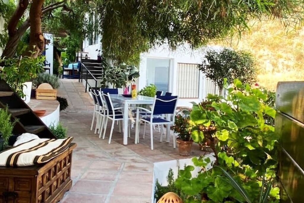 An outdoor patio with a dining table and chairs, surrounded by potted plants and a wooden bench.