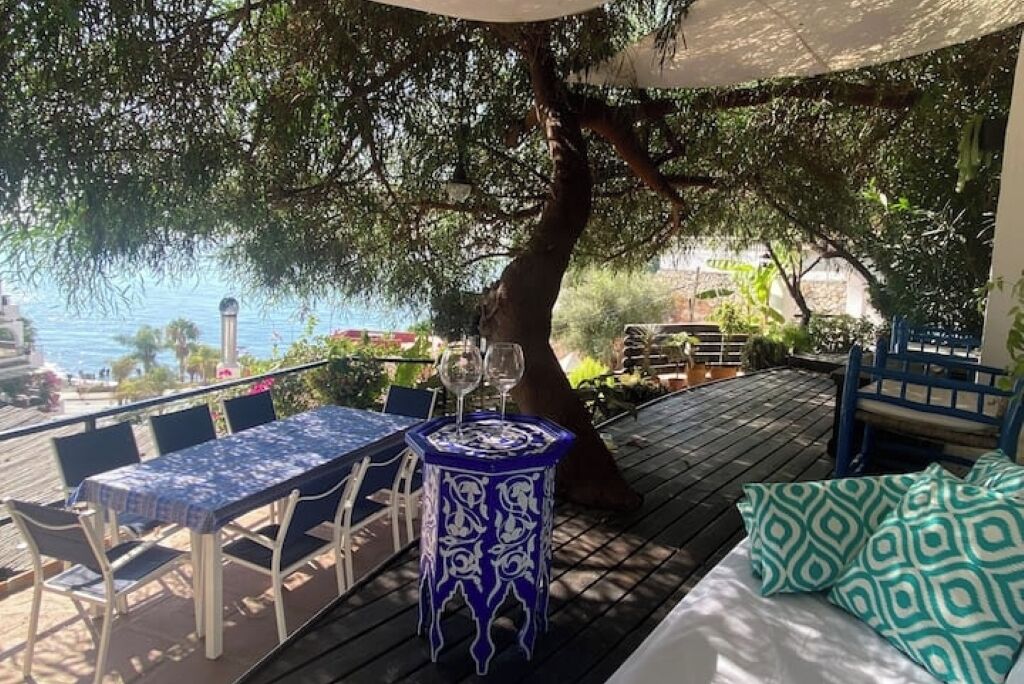 Eine Holzterrasse mit Tisch und Stühlen, einem kleinen Dekorationstisch und einer Bank mit Kissen.