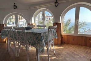 A dining area with a table covered in a floral tablecloth, surrounded by white chairs, large arched windows with a view of the sea, and wooden paneling on the walls. - CASA EVASION at the foot of the beach, incredible views surrounded by a beautiful garden. (Almuñécar)