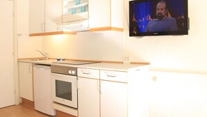 Une cuisine compacte avec des meubles blancs, un four encastré et un four à micro-ondes. Un téléviseur est fixé au mur et diffuse une scène.