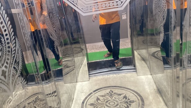 A person standing in a glass elevator with a patterned floor and a green mat at the base. - Vishal Banquet & Marriage Hall (Bodh Gaya)