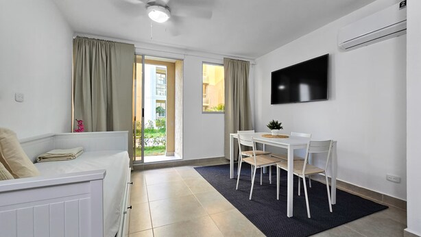 Ett modernt och väl upplyst rum med en takfläkt, en platt-TV som är monterad på väggen, ett matbord med stolar, en soffa och en skjutdörr i glas som leder ut till ett utomhusområde.