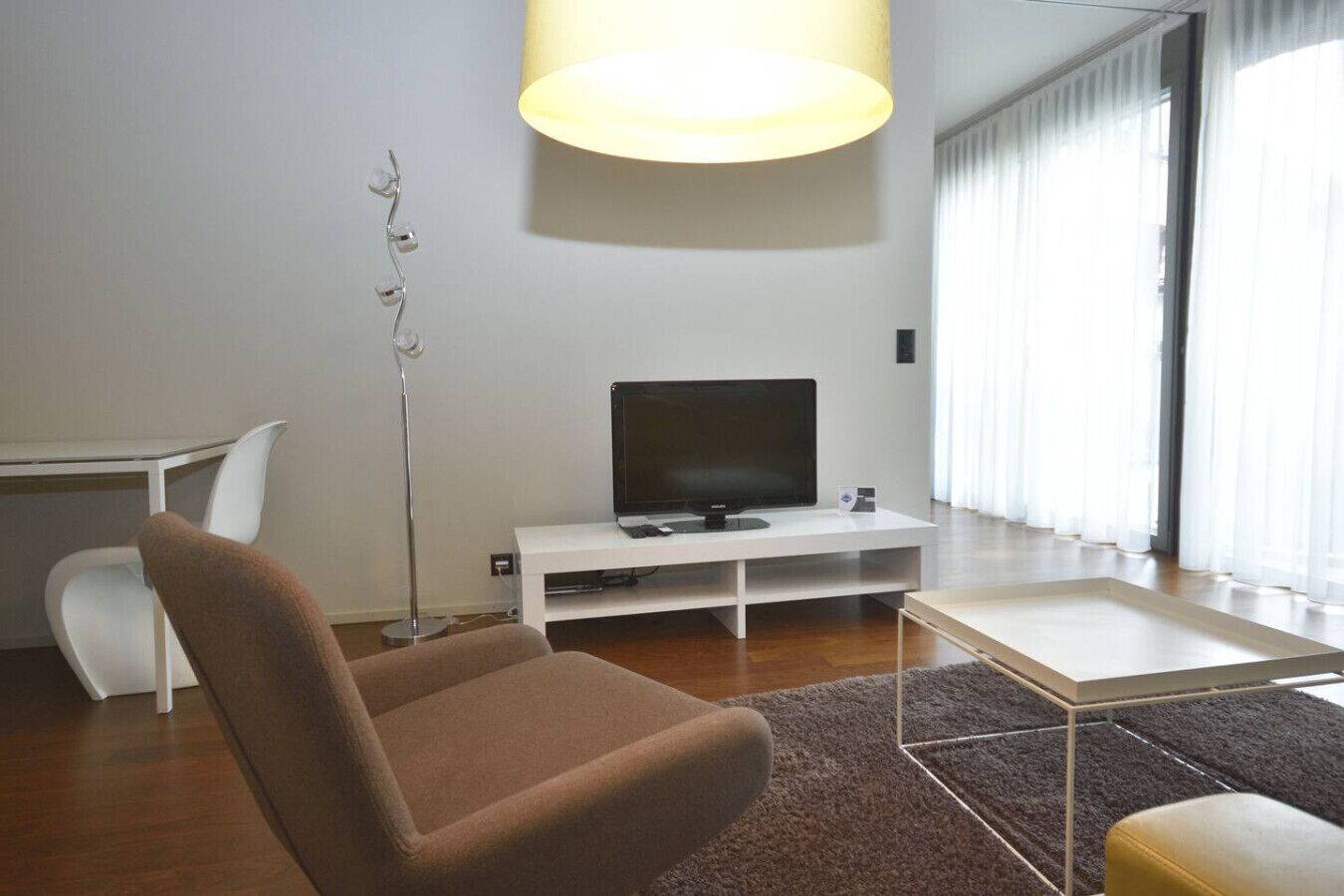 A modern living room with a flat-screen TV on a white stand, a brown armchair, a small white table, and a floor lamp.