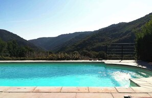 Une piscine à l’eau d’un bleu cristallin, entourée d’une terrasse en pierre, avec en toile de fond de luxuriantes collines verdoyantes et un ciel dégagé.